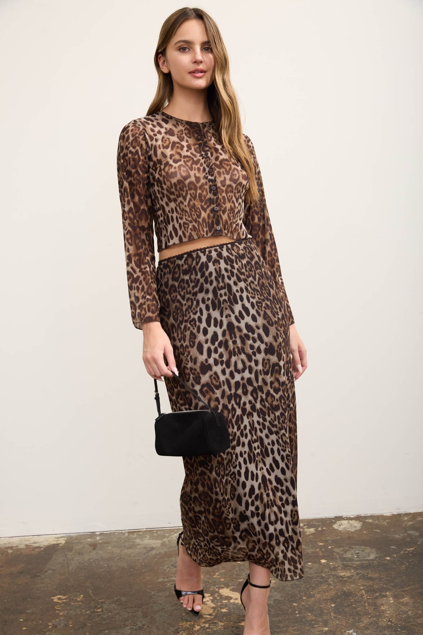 A woman modeling a leopard-print two-piece set consisting of a long-sleeve crop top and a matching midi skirt, paired with black strappy heels and a small black handbag, standing against a plain off-white wall.