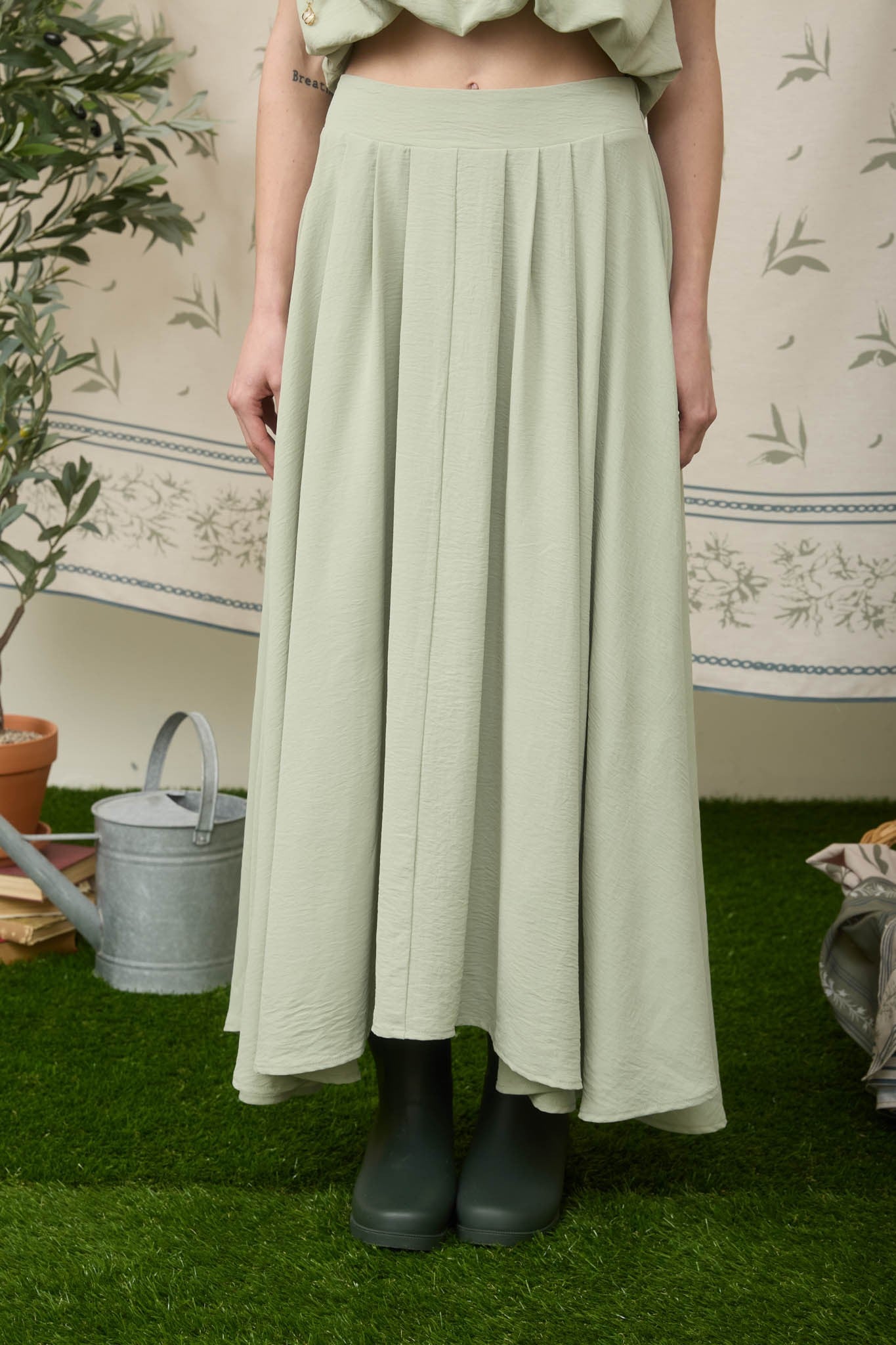 Person wearing a light green pleated skirt standing on grass with a decorative wall in the background.