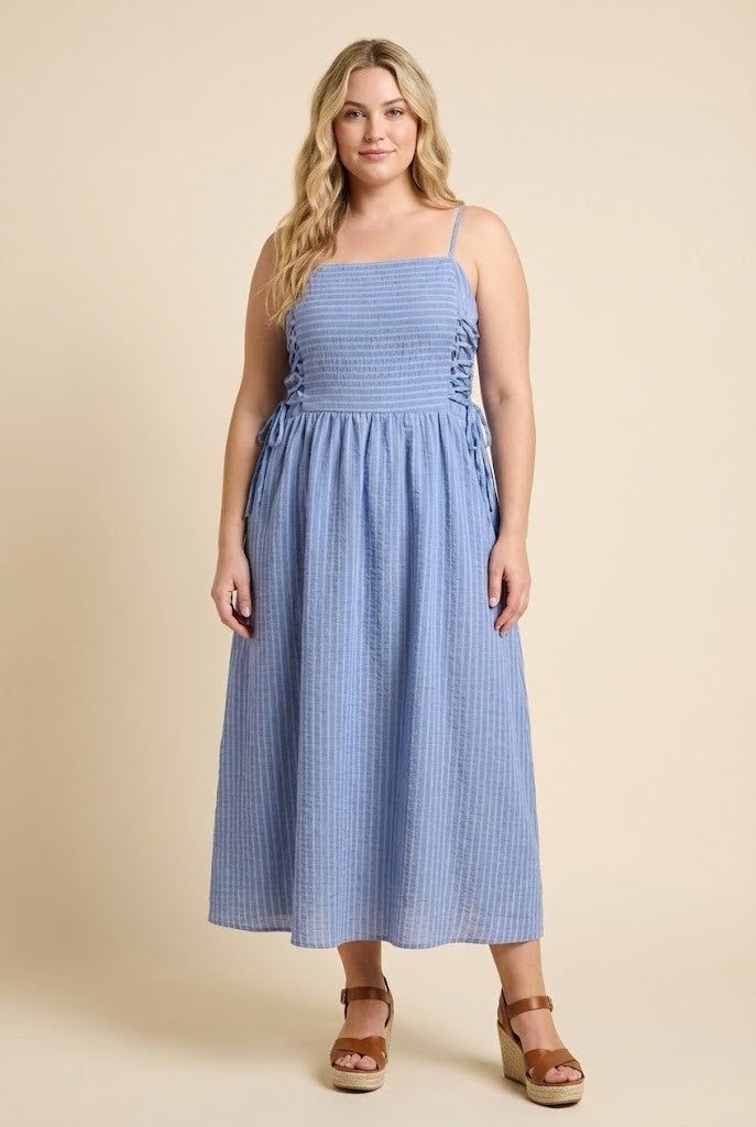 A woman modeling a light blue and white vertically striped midi dress with a smocked bodice and side lace-up details, paired with brown wedge sandals, against a solid beige background.