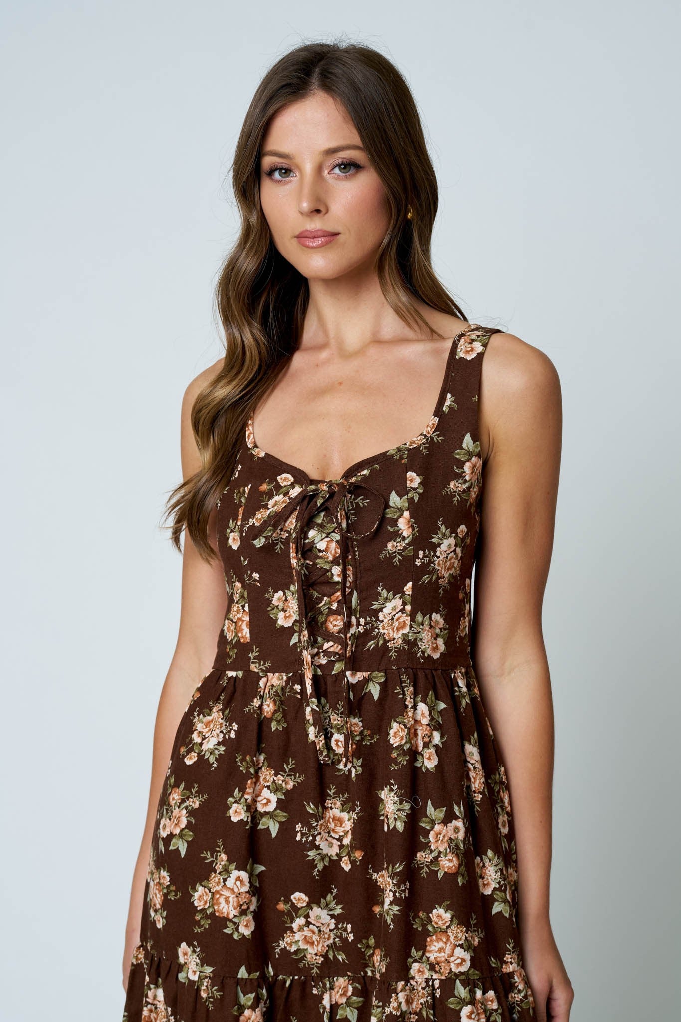 A woman wearing a brown sleeveless dress with a beige and pink floral print, featuring a tie-neck detail and a gathered skirt, photographed against a light gray studio background.