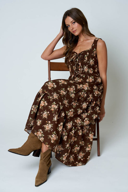 A woman sits on a wooden chair wearing a brown floral midi dress with thin straps and brown ankle boots against a light gray studio background.