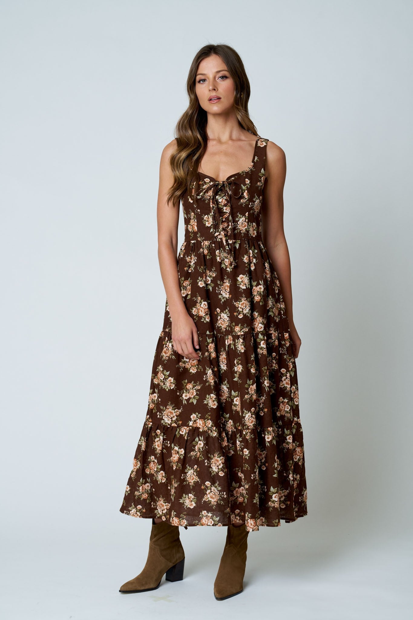 A woman modeling a sleeveless brown midi dress with a beige floral pattern, paired with brown ankle boots, against a light gray studio background.