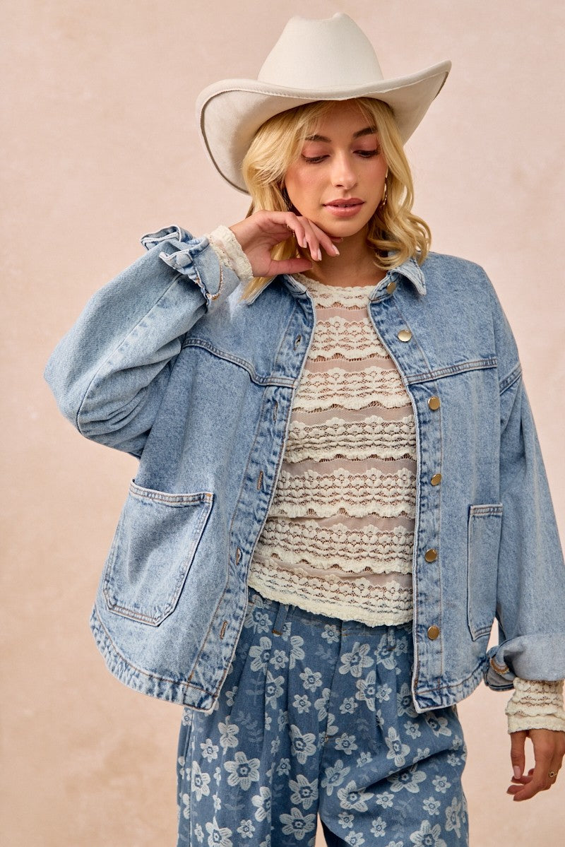 Woman wearing a denim jacket, lace top, and floral pants with a beige hat on a beige background